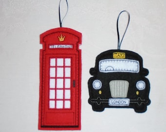 Może przedstawiać: Dwa ozdoby z filcu, jedna to czerwona budka telefoniczna z złotą koroną i napisem "TELEPHONE", a druga to czarna londyńska taksówka z napisem "TAXI" i "LONDON" z przodu.