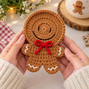 Puede incluir: Un posavasos con forma de hombre de jengibre hecho de hilo de ganchillo marrón. El posavasos presenta una cabeza circular con una sonrisa bordada en negro, una pajarita de hilo rojo brillante en el cuello y bordados en zigzag blanco en las extremidades.