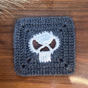 May include: A square crochet granny square featuring a dark charcoal gray border and a central white skull motif. The skull design is created with white yarn stitches against the dark gray background, forming a distinct, centered graphic pattern.