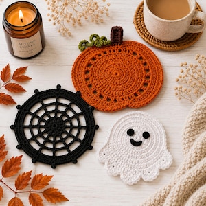 May include: Three crocheted coasters featuring a round orange pumpkin with a brown stem and green vine, a circular black spider web, and a white ghost with black eyes and a smiling mouth, arranged together on a light wooden surface.