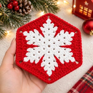 Puede incluir: Un posavasos de ganchillo hexagonal con un motivo de copo de nieve blanco centrado sobre un fondo rojo vibrante. El intrincado trabajo de hilo crea un patrón geométrico de seis puntas, mostrando un diseño de punto limpio y texturizado.