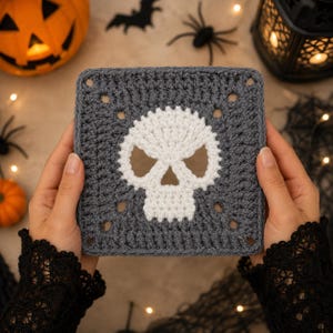 Puede incluir: Una pieza cuadrada de ganchillo que presenta un motivo de calavera blanca centrado sobre un fondo gris oscuro. El diseño está realizado en un estilo clásico de granny square con espacios abiertos en las esquinas y un acabado de hilo texturizado.