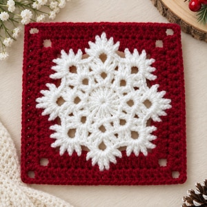 May include: A square crochet motif featuring a deep red border and a central white snowflake pattern. The design consists of intricate white yarn stitches forming a star-like shape against the solid red background, creating a geometric square.