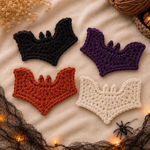 May include: Four crocheted bat appliques in black, purple, burnt orange, and cream. Each bat features a central body with two small ears and wide, scalloped wings. The items are arranged in a grid pattern on a textured, light-colored fabric surface.
