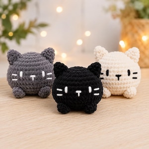 May include: Three round, crocheted cat plushies in gray, black, and cream. Each plush features two small ears, two embroidered eyes, a small nose, and three whiskers on each side. The gray and black cats have white stitching, while the cream cat has black stitching.
