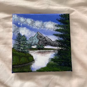 May include: A rectangular canvas painting depicting a landscape scene with a central mountain peak, a reflective body of water in the foreground, and dense green pine trees on the sides. The sky is bright blue with white clouds.