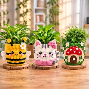 Puede incluir: Tres fundas de ganchillo para macetas que representan una abeja amarilla y negra con alas blancas, un gato crema y rosa con un lazo, y un bosque verde con una casa de setas roja, cada una con una planta verde sobre una base de madera.