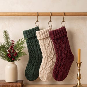 Op de afbeelding: Drie gebreide kerstsokken hangend aan een houten roede. De sokken zijn donkergroen, crème en diep bordeauxrood, elk voorzien van een gestructureerd kabelbreipatroon met een omgeslagen boord aan de bovenkant.