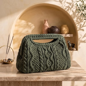 May include: A dark olive green handbag crafted from thick, chunky yarn featuring a prominent cable knit pattern. The bag has a structured rectangular shape with an integrated top handle opening and a textured, woven surface throughout the exterior.