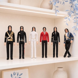 May include: A set of six articulated dolls depicting Michael Jackson in various iconic outfits, including a black military-style jacket with gold accents, a black leather-look suit, a white ensemble, a red cardigan, a black jacket, and a blue jacket.