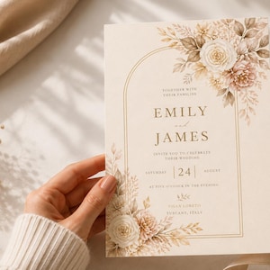 May include: A cream-colored wedding invitation card featuring a gold arched frame, watercolor floral illustrations in beige and brown tones, and the printed text: Together with their families, Emily and James, invite you to celebrate their wedding, Saturday 24 August, at five o'clock in the evening, Villa Loreso, Tuscany, Italy.