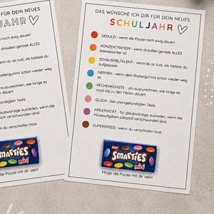 Könnte beinhalten: Zwei identische weiße Karten mit der Überschrift DAS WÜNSCHE ICH DIR FÜR DEIN NEUES SCHULJAHR, einer Liste farbiger Punkte und deutschem Text, jeweils mit einer kleinen blauen Schachtel Smarties Mini, umgeben von verschiedenen Schreibwaren.