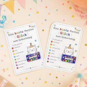 Könnte beinhalten: Zwei rechteckige weiße Karten mit dem Text "Eine bunte Portion Glück zum Geburtstag" und einer Comic-Katze mit Partyhut. Auf jeder Karte ist eine kleine blaue Schachtel Smarties mini befestigt, ergänzt durch eine Liste mit bunten Punkten.