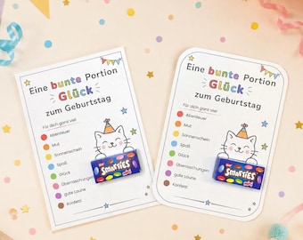 Może przedstawiać: Dwie prostokątne białe karty z napisem "Eine bunte Portion Glück zum Geburtstag" oraz rysunkowym kotem w czapce urodzinowej. Do każdej karty przymocowane jest małe niebieskie pudełko cukierków Smarties mini oraz lista kolorowych punktów.