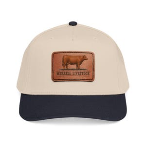 May include: A beige baseball cap with a dark navy blue brim and a top button. Centered on the front is a rectangular brown leather patch featuring an illustration of a brown cow standing on grass with the text MERRELL LIVESTOCK printed below.