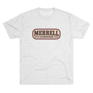 May include: A light heather gray short-sleeve t-shirt featuring a central rectangular graphic with rounded corners. The graphic contains the dark brown text MERRELL LIVESTOCK enclosed within a maroon and white border with horizontal lines.
