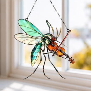 Op de afbeelding: Een glas-in-lood suncatcher in de vorm van een mug die viool speelt. Het insect heeft doorschijnende turquoise en groene vleugels, een gesegmenteerd lichaam met donkere markeringen en een roodachtige kop. Het houdt een oranje-bruine viool vast.