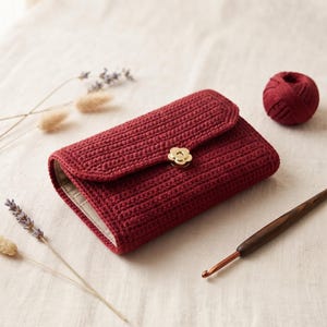 May include: A deep red rectangular crochet wallet featuring a textured stitch pattern and a gold-toned flower-shaped metal clasp. The wallet has a folded flap closure and a light-coloured interior lining visible along the side edge.