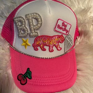 May include: A bright pink and white trucker hat featuring a white front panel decorated with pearl-encrusted letters BP, a pink tiger patch, a diamond patch, a yellow star, and a pink rectangular patch with the text Bad Hair Day. The brim has a cherry patch.