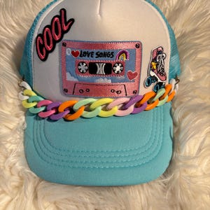 May include: A light blue and white trucker hat featuring a pink cassette tape patch labeled LOVE SONGS, a roller skate patch, and the word COOL in black script. A chunky, multi-colored plastic chain in orange, yellow, and purple accents the brim.