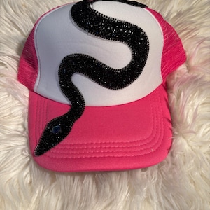 Pode incluir: Um boné trucker rosa choque e branco com um design de cobra sinuosa feito de strass pretos. A cobra estende-se do painel frontal branco até à aba rosa sólida, com um único strass azul a servir de olho para o animal.