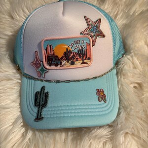 May include: A light blue trucker hat with a white front panel featuring a rectangular patch with a desert landscape, a van, and the text "Take It Easy". The hat is decorated with glittery star, cactus, and flower patches and a gold chain trim.