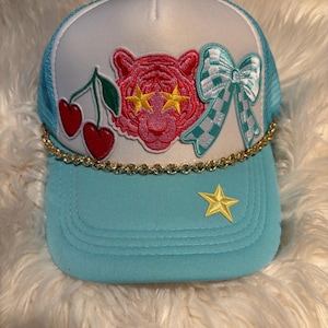 Puede incluir: Una gorra tipo trucker de color azul claro y blanco con una cadena dorada en la base de la parte frontal. El panel delantero presenta parches bordados de cerezas rojas, una cara de tigre rosa con ojos de estrella amarillos y un lazo a cuadros.