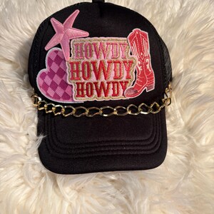 Peut inclure: Une casquette de camionneur noire avec une chaîne dorée sur la visière. Le panneau avant présente un écusson étoile rose, un écusson cœur à carreaux roses et un écusson avec le texte HOWDY en lettres rouges et dorées, ainsi qu'une botte.