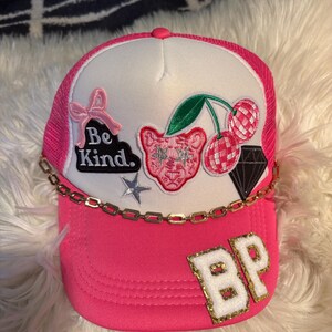 May include: A bright pink and white trucker cap featuring a gold chain across the brim. The front panel displays embroidered patches including a pink bow, a black cloud with the text Be Kind, a pink leopard face, cherries, a diamond, and a star. The letters BP are in white and gold glitter on the brim.