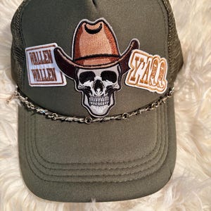 May include: An olive green trucker hat featuring a central embroidered patch of a skull wearing a brown cowboy hat. To the left is a rectangular patch with the text WALLEN WALLEN, and to the right is a patch with the text Y'ALL. A silver-toned barbed wire chain accents the base of the crown.