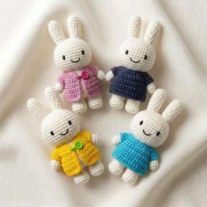 May include: Four crocheted rabbit dolls with white yarn bodies and black embroidered facial features. Each rabbit wears a different colored crocheted garment: a pink cardigan with a pink button, a dark blue dress, a yellow cardigan with a green button, and a blue dress.