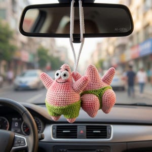 May include: A crochet hanging ornament featuring two pink starfish characters with white eyes and black pupils. One character faces forward with arms raised, while the other faces away. Both wear green shorts, suspended by a white cord from a rearview mirror.