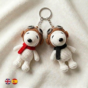 May include: Two white crocheted dog keychains featuring brown aviator caps with goggles. One dog wears a bright red scarf, while the other wears a black scarf. Both figures are attached to a single silver metal split ring by short chains.