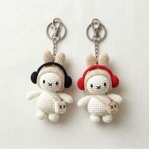 Puede incluir: Dos llaveros de conejo tejidos a ganchillo con cierres de mosquetón de metal. Un conejo lleva auriculares negros y un pequeño bolso beige con una marca X. El otro conejo lleva auriculares rojos y un bolso beige a juego con una marca X.