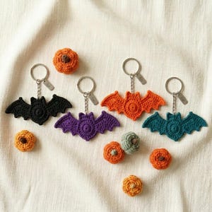 May include: Three crocheted bat keychains in black, purple, orange, and teal, each with a metal ring and chain. Accompanying the bats are several small, round, crocheted pumpkins in shades of orange and green, arranged on a textured fabric.
