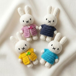 May include: Four crocheted rabbit dolls with white yarn bodies and black embroidered facial features. Each rabbit wears a different coloured cardigan: one pink with a button, one dark blue, one yellow with a green button, and one bright blue.