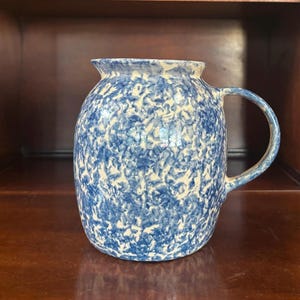 May include: A ceramic pitcher featuring a rounded body and a single handle. The surface is decorated with a blue and white mottled, sponge-painted pattern. The vessel has a wide, slightly flared rim and a smooth, glossy finish throughout the exterior.