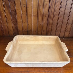 May include: A rectangular, off-white ceramic baking dish with integrated side handles. The dish features a smooth, matte interior surface and a slightly weathered, neutral-toned exterior finish, designed with a wide, open basin for food preparation.