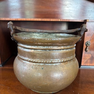 May include: A round copper pot with a weathered patina and a wide, open top. The vessel features a thick, dark, rusted iron swing handle attached to two side lugs. A decorative rope-like embossed band encircles the upper body of the pot.