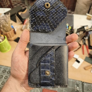 Op de afbeelding: Een verticale lederen portemonnee met een donkerblauwe klep met slangenleertextuur en een koperen drukknoopsluiting. De behuizing is gemaakt van glad zwart leer met een bijpassende blauwe slangenleren accentstrook met een drukknoop.