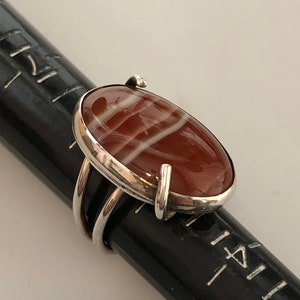 Pu&ograve; includere: Un anello d'argento con una pietra di agata rossa e bianca. La pietra &egrave; di forma ovale e ha una finitura lucida. L'anello &egrave; posto su una superficie nera.