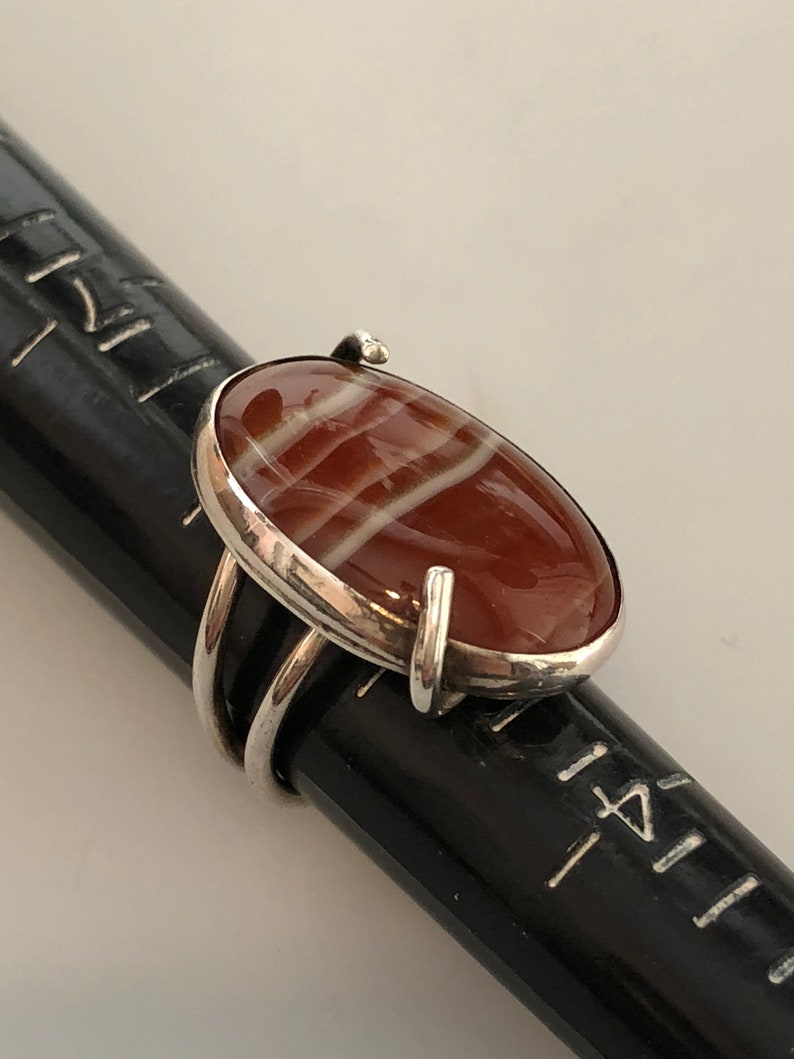 Pu&ograve; includere: Un anello d'argento con una pietra di agata rossa e bianca. La pietra &egrave; di forma ovale e ha una finitura lucida. L'anello &egrave; posto su una superficie nera.