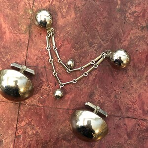 May include: A set of gold-toned cufflinks with a chain and three round balls. The cufflinks are made of a smooth, shiny metal.