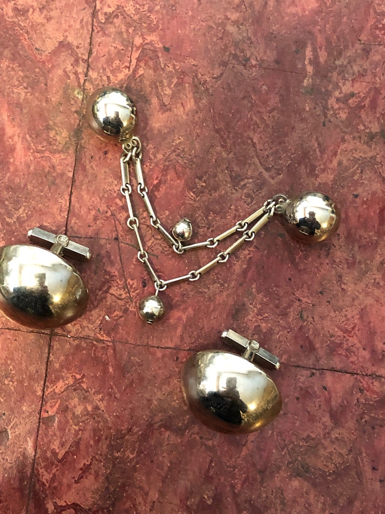 May include: A set of gold-toned cufflinks with a chain and three round balls. The cufflinks are made of a smooth, shiny metal.