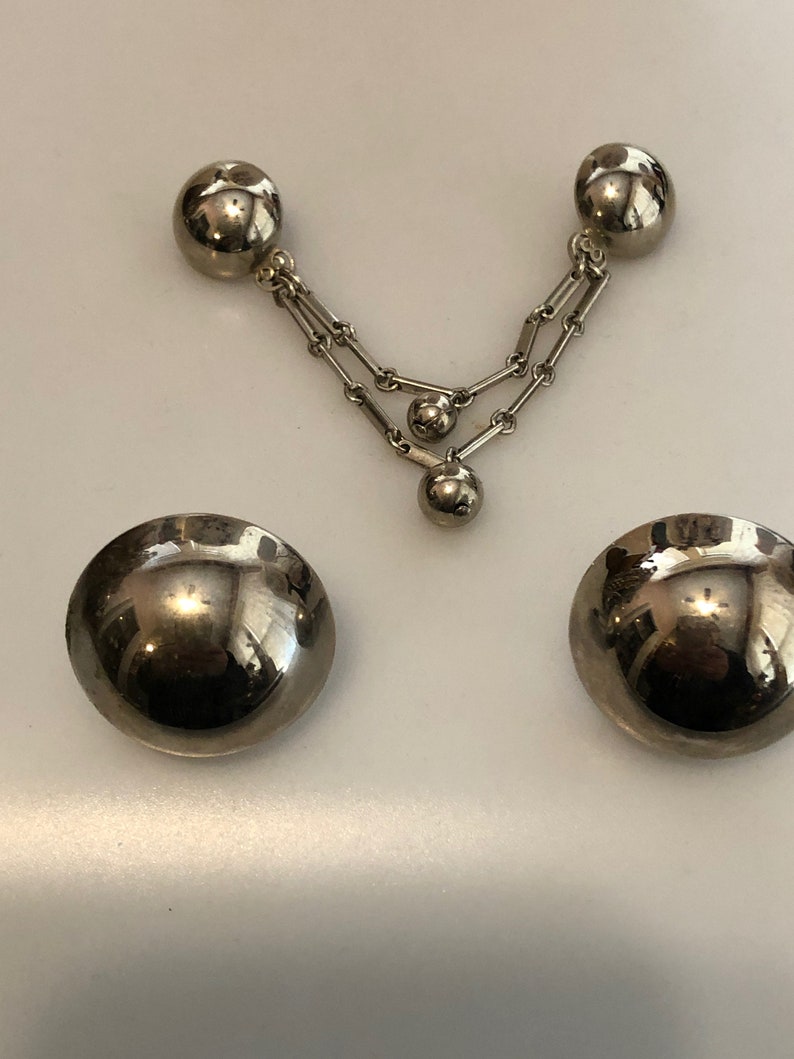 May include: A pair of silver tone clip-on earrings with a dangling chain and three round beads. The earrings are made of metal and have a shiny finish.