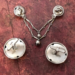 May include: A set of silver cufflinks with a chain and a dangling ball. The cufflinks have a circular design with a small crossbar on the top.