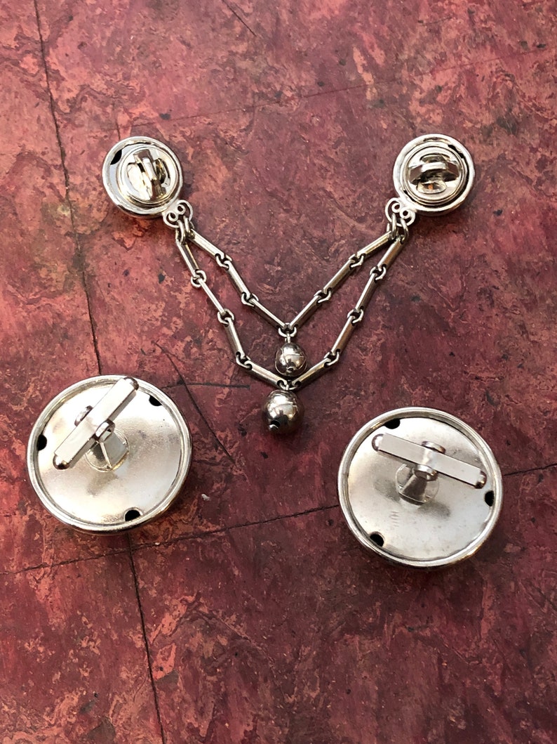 May include: A set of silver cufflinks with a chain and a dangling ball. The cufflinks have a circular design with a small crossbar on the top.