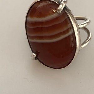 Pu&ograve; includere: Un anello d'argento con una grande pietra di corniola di forma ovale. La pietra ha delle strisce bianche che la attraversano.