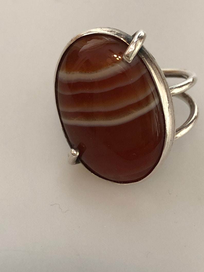 Pu&ograve; includere: Un anello d'argento con una grande pietra di corniola di forma ovale. La pietra ha delle strisce bianche che la attraversano.