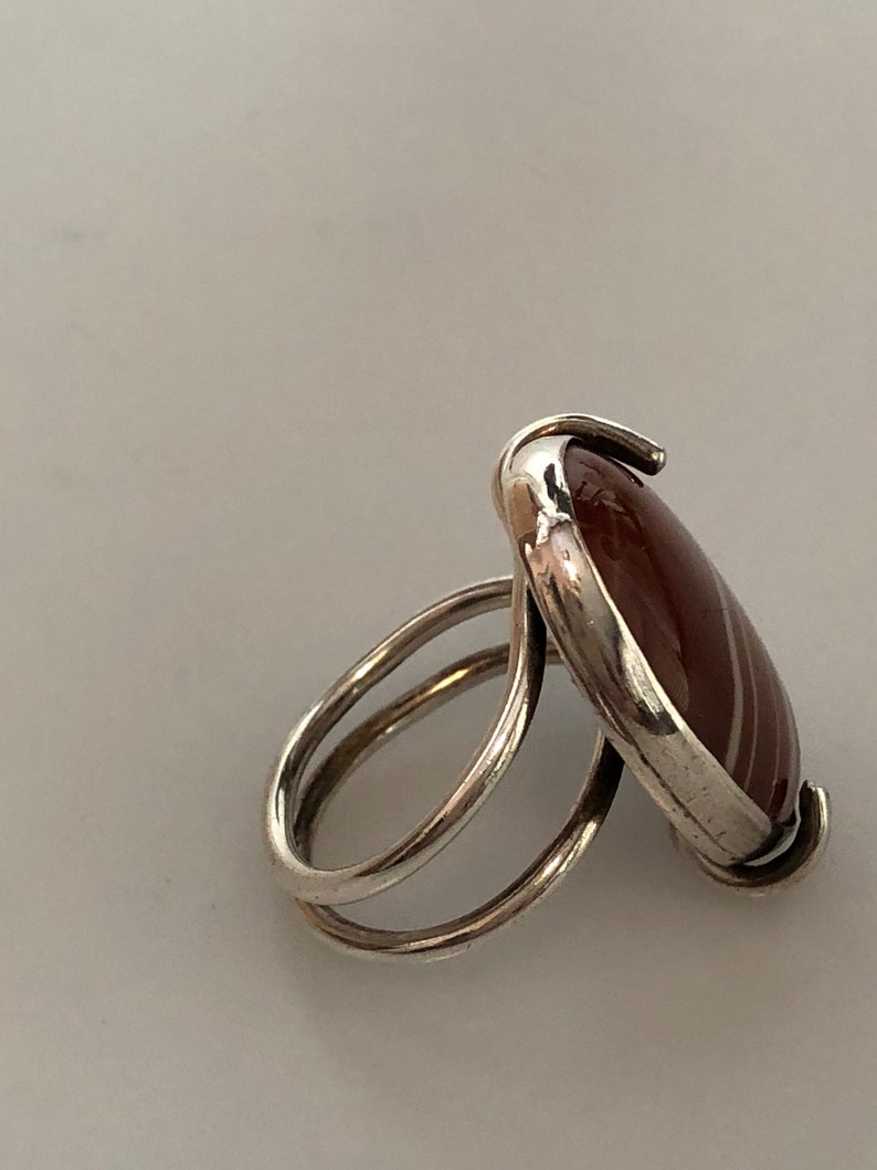Pu&ograve; includere: Un anello d'argento con una pietra di agata rossa e bianca incastonata in una montatura a castone. L'anello ha una doppia fascia.